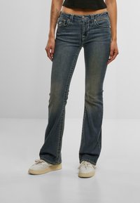 Razširjene traper hlače v zbledeli modri barvi z kontrastnim šivom, petimi žepi in gumbnim zapiranjem. Kombinirane z belimi supergami.