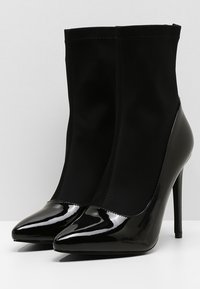 Bottes noires à talons hauts et bout pointu, alliant tissu mat sur la tige et cuir verni brillant sur la partie inférieure et le talon.