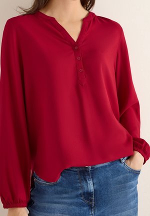 Femme portant un chemisier rouge à manches longues avec des boutons au niveau de l'encolure et un jean bleu en denim, main dans la poche.