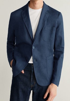 Uomo che indossa un blazer blu navy sopra una t-shirt bianca e jeans blu scuro, con una mano in tasca.