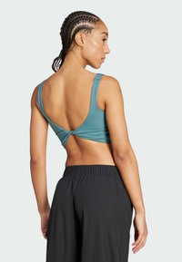 Teal sportbeha met brede bandjes en een gedraaide achterkant. Gecombineerd met zwarte, losse broek. Gladde textuur, minimalistische stijl.