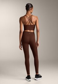 Hnědý sportovn�í crop top s křížovými popruhy, doplněný o odpovídající legíny s vysokým pasem. Černé sportovní boty s bílou podrážkou.