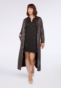 Donna con un lungo cappotto di pelle marrone scuro sopra un vestito a stampa leopardata nera, tacchi beige a punta, che cammina su uno sfondo chiaro e semplice.
