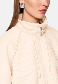 Beige zip-up jacket with textured stripes, high collar, and dual drawstrings; paired with a large, reflective geometric earring.