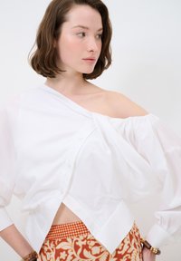 Blouse blanche, décolletée épaules nues, à coupe ample, avec une fermeture par boutons à l'avant et des manches froncées. Associée à un pantalon orange à motifs.
