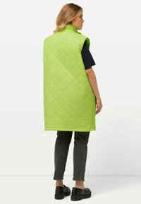Grüne gesteppte ärmellose Weste mit hohem Kragen und abgerundetem Saum, getragen über einem marineblauen Shirt. Kombiniert mit schwarzen Schuhen mit dicker Sohle.
