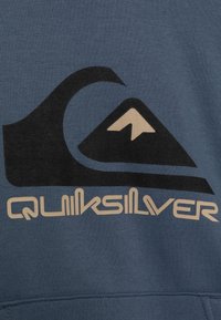 Modrá mikina s velkým černým a krémovým logem Quiksilver, které obsahuje design hory a vlny. Vyrobeno z měkkého materiálu s texturovaným povrchem.