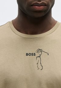 Κοντινό πλάνο ενός ατόμου που φοράει μπεζ πουκάμισο με το κείμενο "BOSS" και μια μινιμαλιστική φιγούρα γκολφέρ να κουνάει ένα κλαμπ τυπωμένα στο στήθος.