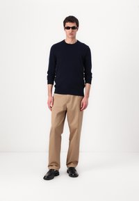 Pull marin, pantalon beige et chaussures noires. Le mannequin porte des lunettes de soleil minces et rectangulaires. Design simple avec des couleurs unies et une silhouette ajustée.