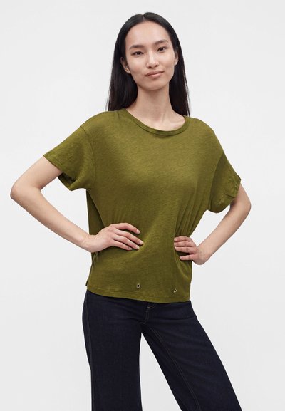Femme aux longs cheveux foncés debout, les mains sur les hanches, portant un haut à manches courtes vert olive et un jean foncé sur un fond uni.