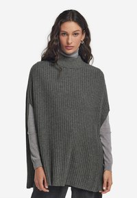 Grå, ribbad stickad poncho med hög krage, avslappnad passform och sidöppningar. Bärs över en långärmad, ljusgrå topp och mörka jeans.