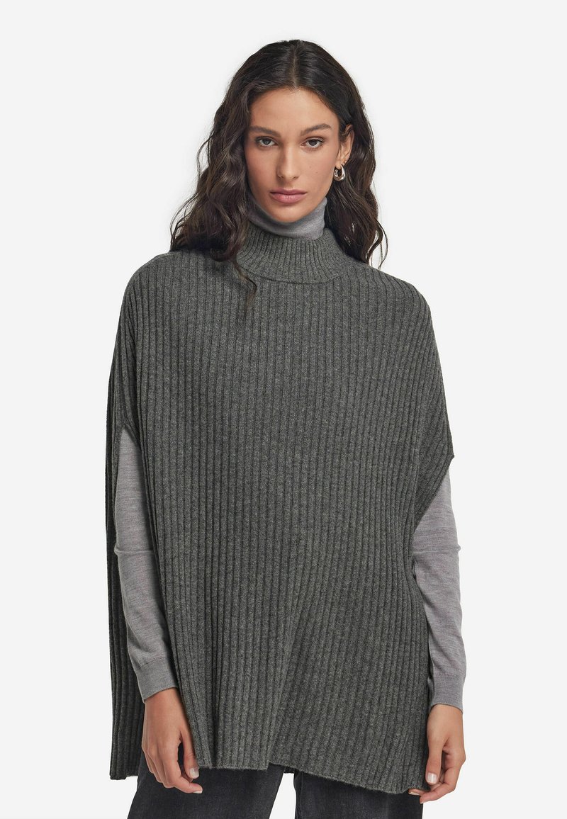 Grå, ribbad stickad poncho med hög krage, avslappnad passform och sidöppningar. Bärs över en långärmad, ljusgrå topp och mörka jeans.