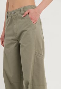Pantalons larges en coton vert avec une poche avant, coupe droite et détails de couture visibles sur l'ourlet et le côté.