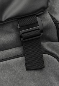 Der graue Stoff-Rucksack hat einen schwarzen Plastikverschluss und Riemen, mit einer glatten Textur auf der Klappe und einem kontrastierenden strukturierten Körper.