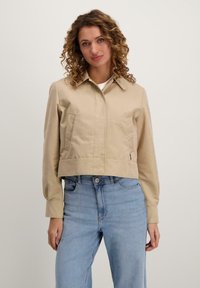 Beige cropped jack met een kraag, twee voorzakken en een gladde textuur. Gecombineerd met lichtblauwe jeans.