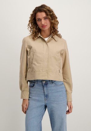 Beige cropped jack met een kraag, twee voorzakken en een gladde textuur. Gecombineerd met lichtblauwe jeans.