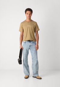 Tan cotton t-shirt with white logo, blue denim jeans, beige slip-on shoes, and a black shoulder bag held in left hand. Casual fit.