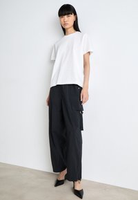 T-shirt blanche à manches courtes associée à un pantalon cargo noir ample doté de poches et d'accents en métal doré, complété par des chaussures noires à bout pointu.