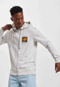 Lys grå hettegenser med glidelås laget av mykt materiale, med en frontlogo i oransje aksenter. Designet inkluderer en hette med snor og ribbestrikkede mansjetter.