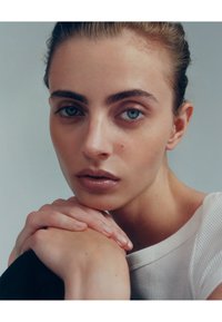 Portræt af en person med lys hud, blå øjne og brunt hår, iført en hvid ribbed top, mod en blød grå baggrund. Hænderne hviler på en arm.