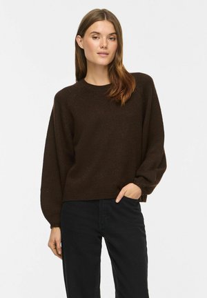 Femme aux longs cheveux bruns portant un pull marron foncé à manches longues et un pantalon noir, debout avec une main dans la poche sur fond blanc.