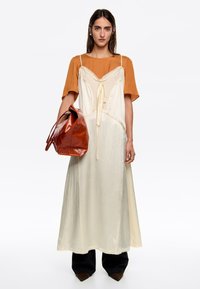 Kremowa sukienka maxi z koronkowym wykończeniem, zestawiona z pomarańczowym topem z krótkim rękawem oraz błyszczącą czerwoną skórzaną torebką. Czarne spodnie o szerokich nogawkach.