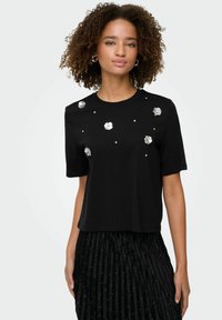 Sort kortærmet t-shirt med blomsterdekorationer og små perler som detalje. Blødt stof med en afslappet pasform og rund halsudskæring.