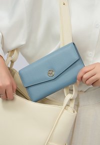 Portafoglio in pelle testurizzata azzurra con dettaglio logo oro, parzialmente visibile in una borsa color crema con un design semplice e liscio.