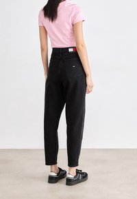 Jeans negros de cintura alta con un ajuste relajado, con bolsillos traseros y un parche con el logo en la cintura. Combinados con zapatillas negras y una blusa rosa.