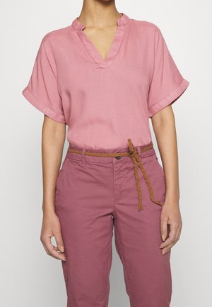 Person trägt eine kurzärmelige rosa Bluse, die in pinke Hosen gesteckt ist, mit einem geflochtenen braunen Gürtel, der in der Taille gebunden ist, und steht vor einem schlichten Hintergrund.
