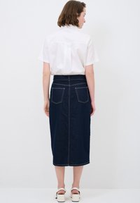 Camisa blanca de manga corta combinada con una falda midi de denim oscura que presenta dos bolsillos traseros y costuras en contraste. Sandalias de tacón cuadrado visibles.