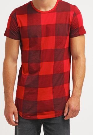 Mężczyzna w czerwono-ciemnoczerwonej, kraciastej koszulce z krótkim rękawem, stojący z luźno opuszczonymi ramionami.