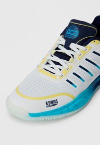 Chaussure de padel K-Swiss blanche avec des accents navy et jaune, semelle bleue, lacets blancs et logo de la marque sur la languette et le côté.