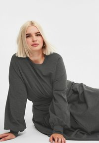 Młoda kobieta z blond bobem, ubrana w długą szaro-ciemną sukienkę, pochylona do przodu, z dłońmi na podłodze, na białym tle.