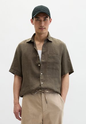 Man draagt een donkergroen overhemd met korte mouwen en knopen, beige broek met trekkoord, wit ondershirt en zwarte pet, staand voor een effen achtergrond.