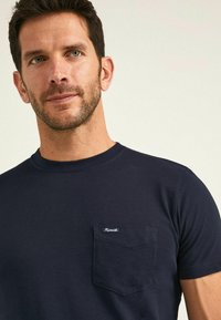 Camiseta de algodón azul marino con cuello redondo, mangas cortas y un bolsillo en el lado izquierdo del pecho que presenta una pequeña etiqueta de la marca. Textura suave.