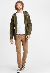 Green zip-up hoodie with textured fabric, white T-shirt underneath, brown corduroy pants, and navy sneakers, featuring tassels on hoodie drawstrings.