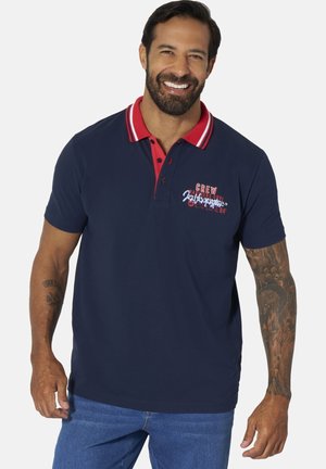 Lächelnder Mann, der ein marineblaues Polo mit rotem Kragen und grafischem Aufdruck, blaue Jeans und sichtbare, detaillierte Tattoos am linken Arm trägt.