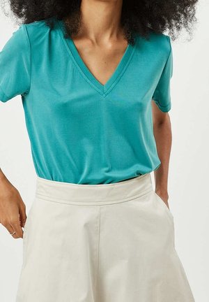 Kvinde iført en teal V-hals kortærmet top, der er stukket ind i en beige nederdel med høj talje og lommer, hænderne nær lommerne, krøllet hår synligt.