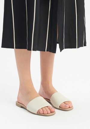 Sandali slide beige chiaro con una larga fascia piatta e una suola bassa color tan. Il design è minimalista e liscio, senza motivi né abbellimenti.