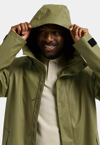Oliven grøn vandtæt jakke med hætte, lynlåslukning foran, trykknapper og sorte Velcro-accenter. Har glat, skinnende tekstur.