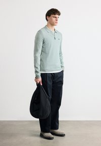 Νεαρός άνδρας στέκεται κρατώντας μια μαύρη τσάντα, φοράει ένα ανοιχτό πράσινο πουκάμισο μακριάς μανίκας, μαύρο παντελόνι και ανοιχτό γκρι μπότες σε απλό φόντο.