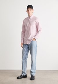 Jonge man die een zwarte pet, lichtroze overhemd met knopen, lichtblauwe jeans en zwarte schoenen draagt, die op een betonnen vloer staat tegen een witte muur.