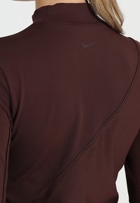 Maronin värinen pitkähiha toppi, jossa on korkea kaulus, ribattu pinta ja huomaamaton musta Nike-logo takana. Sisältää kaarevan sauman yksityiskohtia.