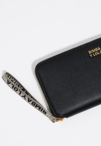 Cartera de cuero negro con superficie texturizada, detalles en cremallera dorada y una correa de tela para la muñeca con un patrón de logo.