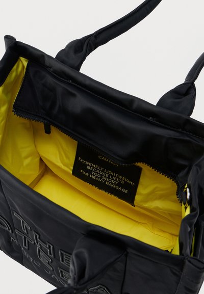 Μαύρη τσάντα ώμου με κίτρινη εσωτερική επένδυση που εμφανίζει μια ετικέτα προειδοποίησης σχετικά με τον ελαφρύ σχεδιασμό και τις κοντές χειρολαβές για βαριά φορτία.