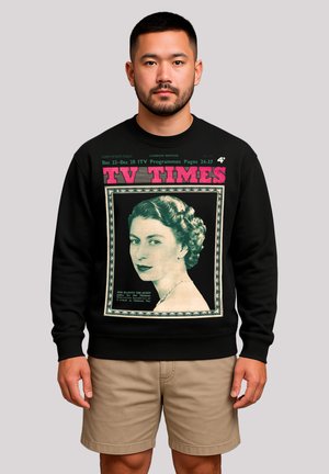 Mann trägt einen schwarzen Sweatshirt mit einem Cover der Zeitschrift TV Times, das ein Vintage-Porträt einer Frau zeigt, umrahmt von Text und einem dekorativen Rand.