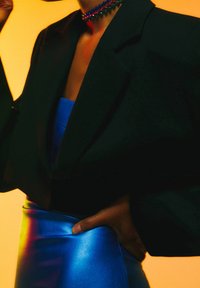 Personne portant un blazer noir, une robe en satin bleue et un collier de perles coloré sur un fond jaune, main posée sur la hanche.