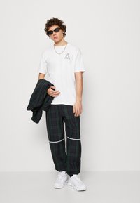 Camiseta blanca con un gráfico de triángulo, pantalones de cuadros negros con rayas blancas contrastantes y zapatillas deportivas blancas. Gafas de sol y una chaqueta negra.