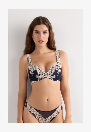 Vrouw met lang bruin haar, gekleed in een marineblauw lingeriesetje met beige bloemenkant details, staande tegen een effen lichte muur.